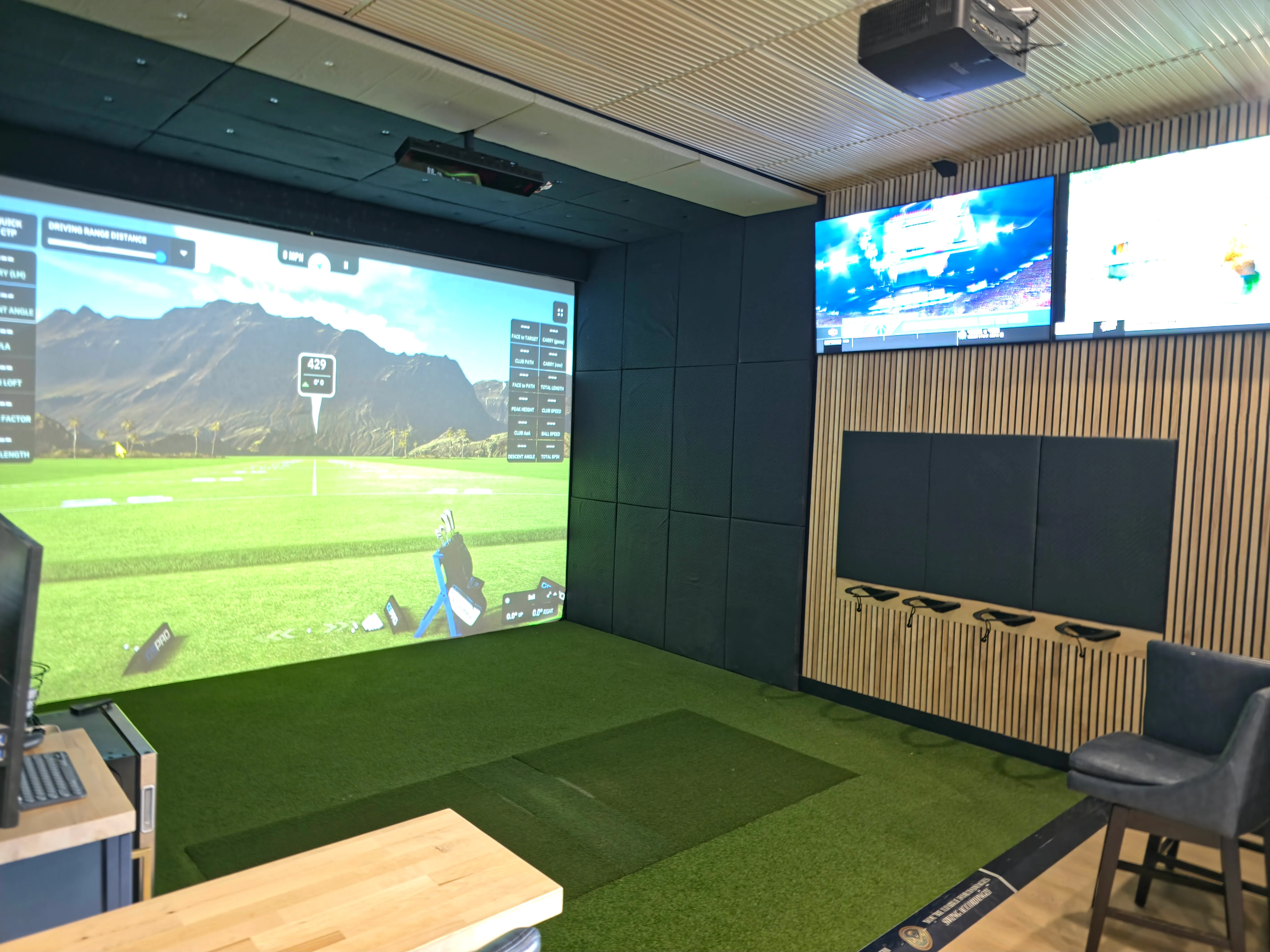Finished golf simulator room with turf, a club rack on a wood-slat wall, and lounge seating — a build like those All Seasons creates for Lake homes, shown at the Kansas City showroom