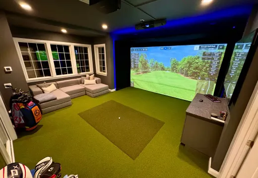 Clean finished home golf simulator room with sectional seating, turf, and accent lighting