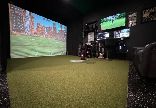 Cluttered home golf simulator room with a desert course on screen and gear along the walls