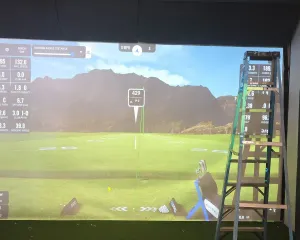 Installer squaring and tensioning the impact screen with a ladder in a golf simulator bay